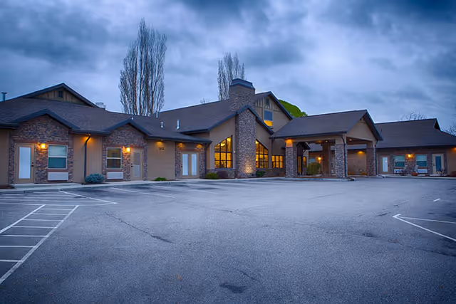 Exterior view of a single-story assisted living facility building at dusk with stone and beige walls, multiple windows, a covered entrance, and a large empty parking lot in front under a cloudy sky.