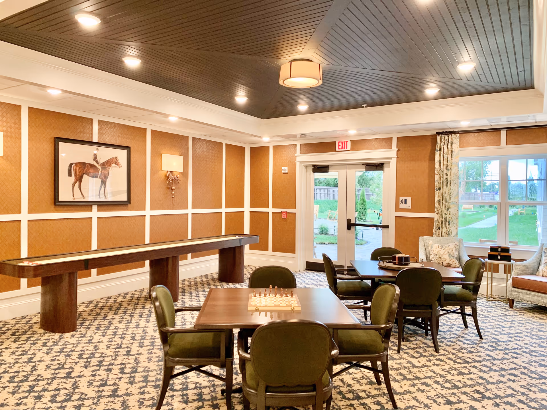 A well-lit common room with patterned carpet, several tables and green cushioned chairs arranged for games, including a chessboard on one table. There is a long shuffleboard table against the wall, a framed picture of a horse and rider, wall sconces, and a ceiling light fixture. Double glass doors and large windows provide a view of a green outdoor area.