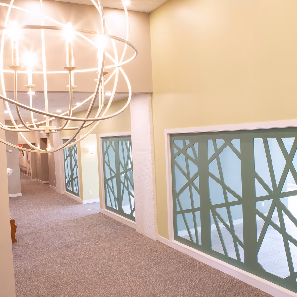 Interior hallway of a senior living facility with beige walls and carpeted floor. Large decorative windows with green geometric patterns line one side of the hallway. A modern spherical chandelier with multiple lights hangs from the ceiling.