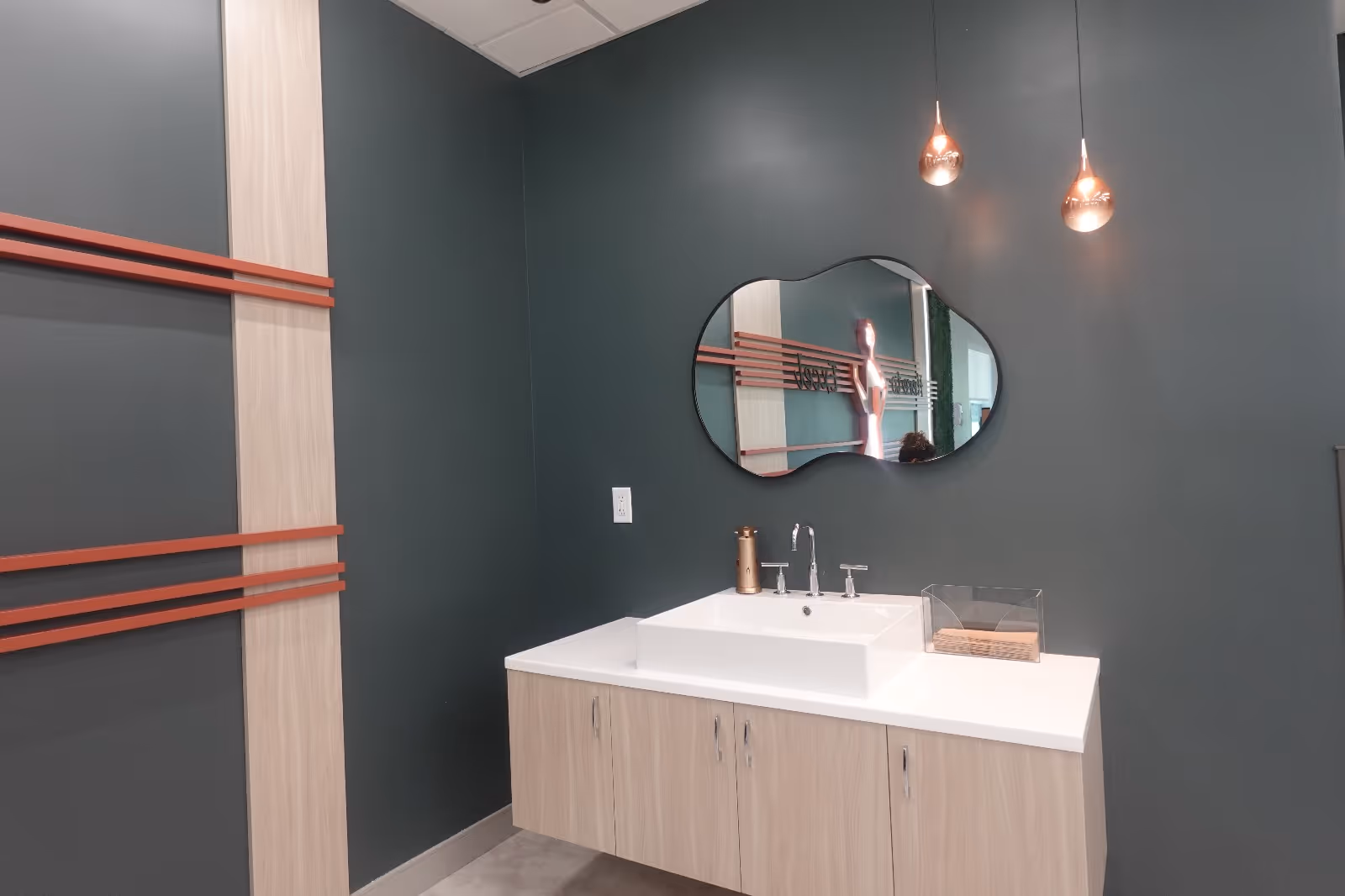 Modern bathroom vanity with a rectangular sink, irregular mirror, pendant lights and light wood cabinets against a dark teal wall.