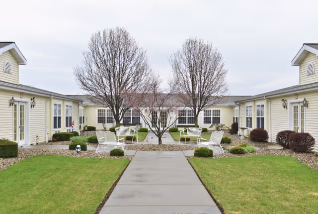 Outdoor courtyard area of a senior living facility with a concrete walkway leading to a seating area with white metal chairs and benches. The courtyard is surrounded by single-story buildings with light yellow siding and multiple windows. There are leafless trees and small bushes around the seating area and along the buildings.