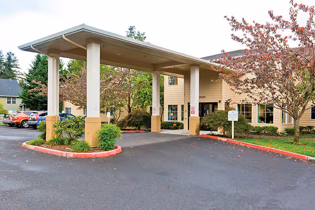 Entrance of Brookdale Fisher's Landing facility showing a covered drop-off area with beige pillars and roof. Surrounding the entrance are trees with autumn foliage and a parking lot with several cars visible on the left side.