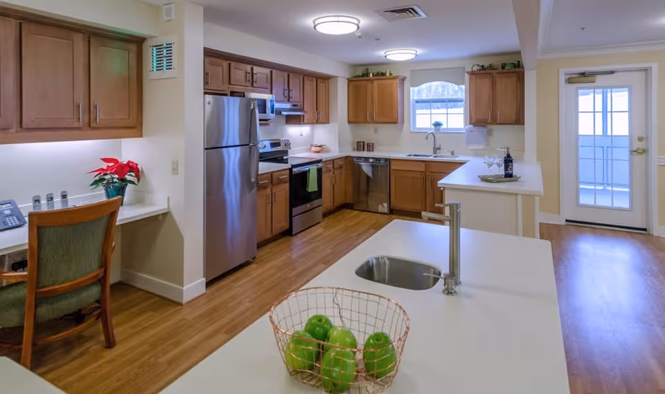 A modern kitchen with wooden cabinets, stainless steel refrigerator, stove, and dishwasher. The kitchen features a white countertop island with a built-in sink and a wire basket containing green apples. There is a small desk area with a chair and a poinsettia plant on the left side. A door with glass panels is visible on the right side, allowing natural light into the room.