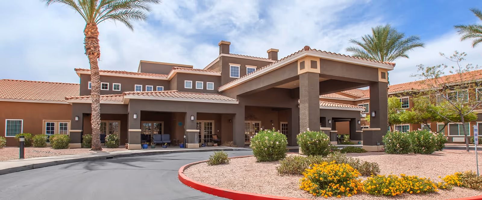 Exterior view of Brookdale Chandler Regional senior living facility showing a covered entrance with columns, desert landscaping with palm trees and flowering bushes, and a clear blue sky.