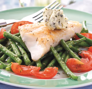A plated meal featuring a piece of cooked white fish topped with a dollop of herb butter, served on a bed of green beans and sliced tomatoes on a green-rimmed plate with a fork in the background.