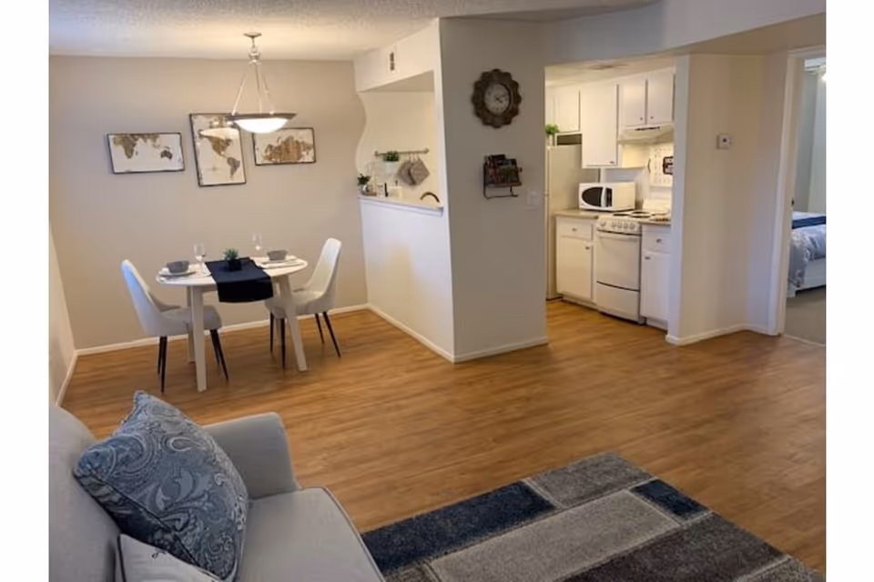 View of a small dining area with a round table set for two, adjacent to a compact kitchen with white cabinets and appliances. The room has wooden flooring and light-colored walls. A gray sofa with patterned cushions and a blue and gray rug are partially visible in the foreground. A bedroom is visible through an open doorway to the right.