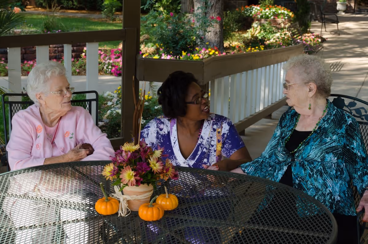 Three elderly women sitting around a metal outdoor table with a flower arrangement and small pumpkins, engaged in conversation on a covered patio with garden flowers and greenery in the background.
