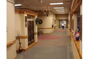 Carpeted interior hallway of a retirement community with an elevator, handrails, seating and a person walking in the distance.