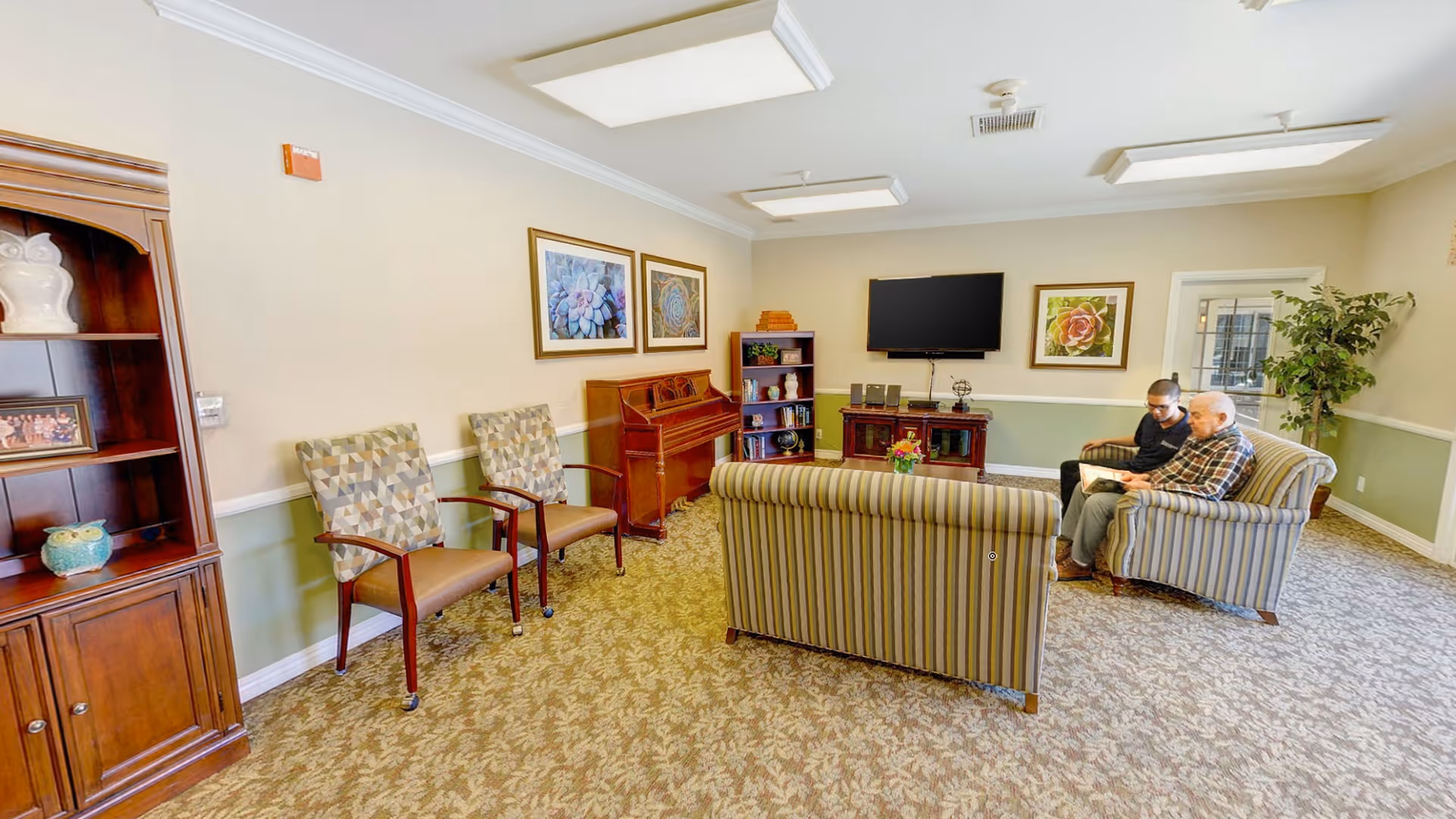 A cozy living room in Silverado Tustin Hacienda Memory Care Community with patterned carpet, two striped sofas, two patterned chairs, a wooden bookshelf, a piano, a TV mounted on the wall, and two people sitting on one sofa looking at a book. The walls are painted beige and green with framed artwork and a potted plant near a glass door.
