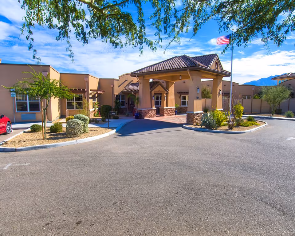 Exterior view of Canyon Valley Memory Care Residence showing a single-story building with a covered entrance, desert landscaping with small trees and shrubs, a flagpole with an American flag, and a clear blue sky with some clouds.