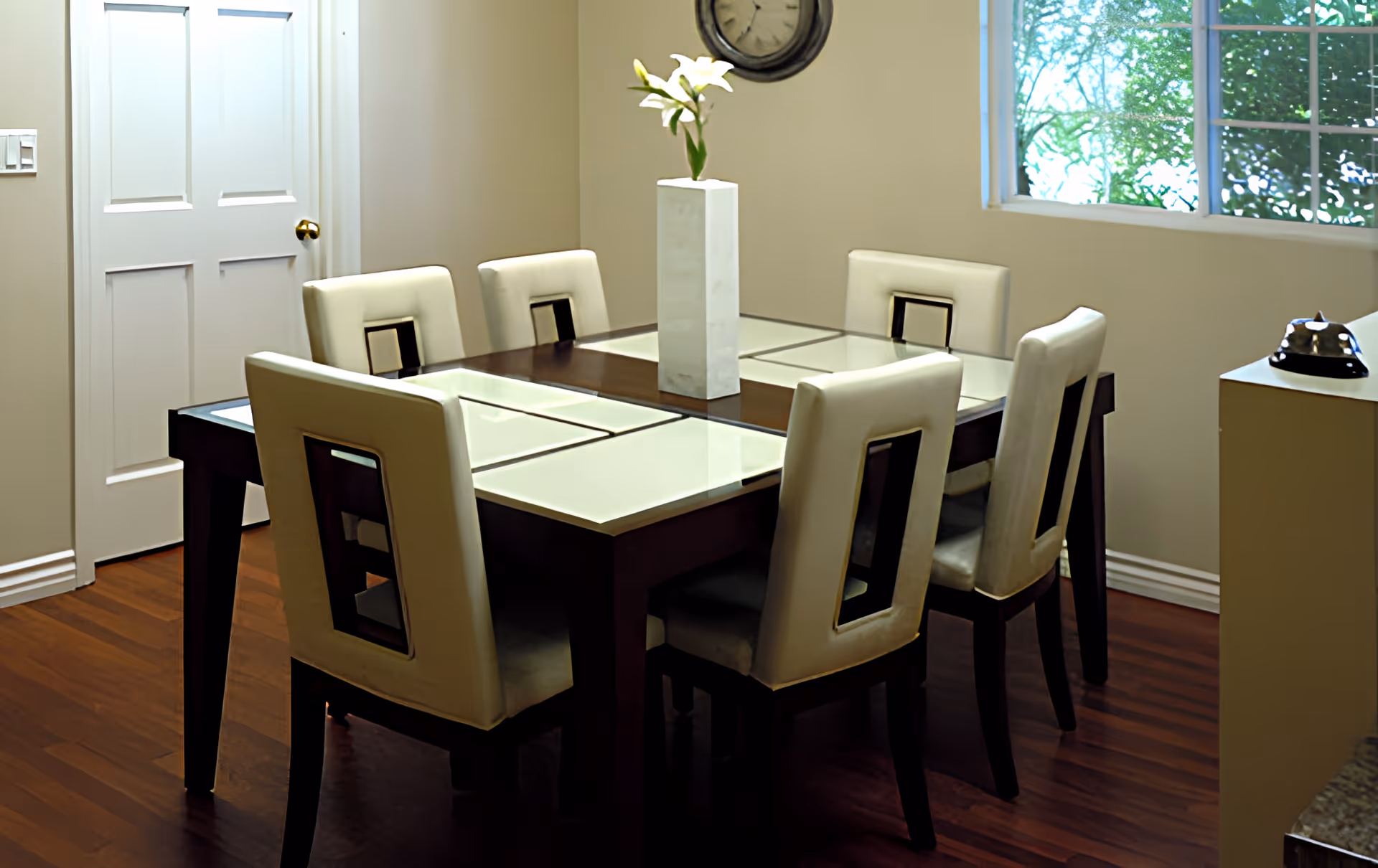 A dining room with a dark wooden table featuring glass panels and six cream-colored chairs with rectangular cutouts in the backrests. A tall white vase with white flowers is placed in the center of the table. The room has wooden flooring, a white door, a wall clock, and a window showing greenery outside. A small table with a silver bell is visible on the right side.