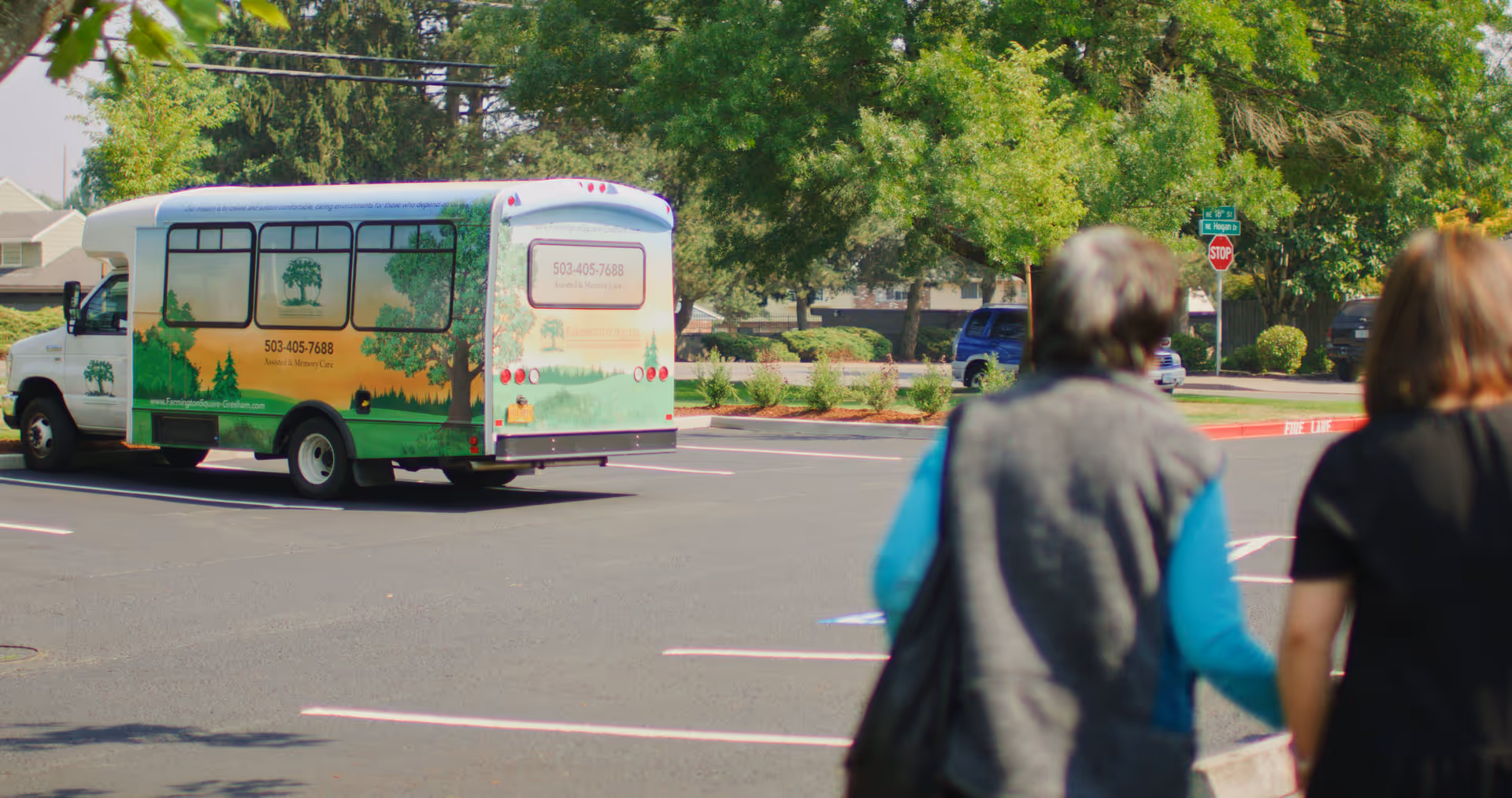 A parking lot with a white and green shuttle bus parked. The bus has signage for Farmington Square at Gresham, including a phone number and website. Two people are walking away from the camera towards the bus. Trees and street signs are visible in the background.