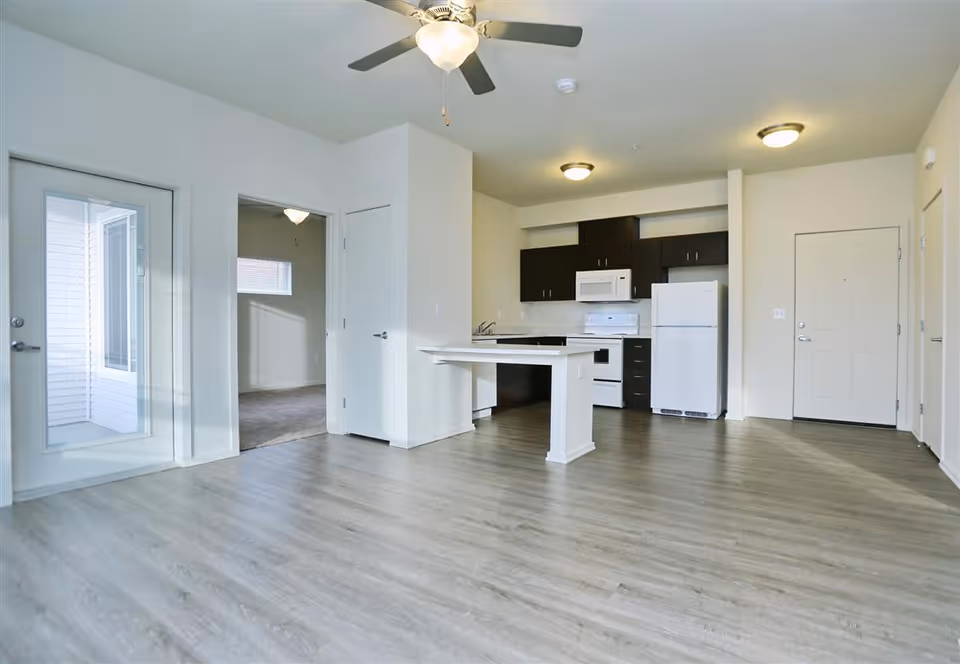 An empty modern apartment interior featuring a kitchen with white appliances including a refrigerator, stove, and microwave, dark cabinetry, and a small island with a white countertop. The room has light wood flooring, white walls, a ceiling fan with light, and a door leading to another room with carpet. There is also a glass door leading outside or to a balcony.