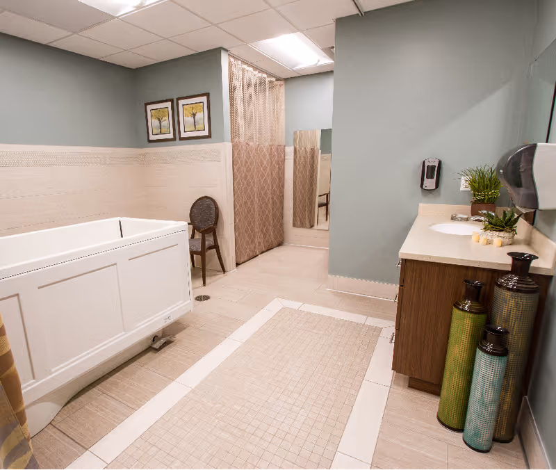 A therapy room with a white therapy tub on the left, a wooden chair against the wall, a beige privacy curtain, a large mirror, and a wooden vanity with a sink on the right. The walls are painted light green and decorated with two framed pictures of trees. There are three decorative tall vases on the floor near the vanity.