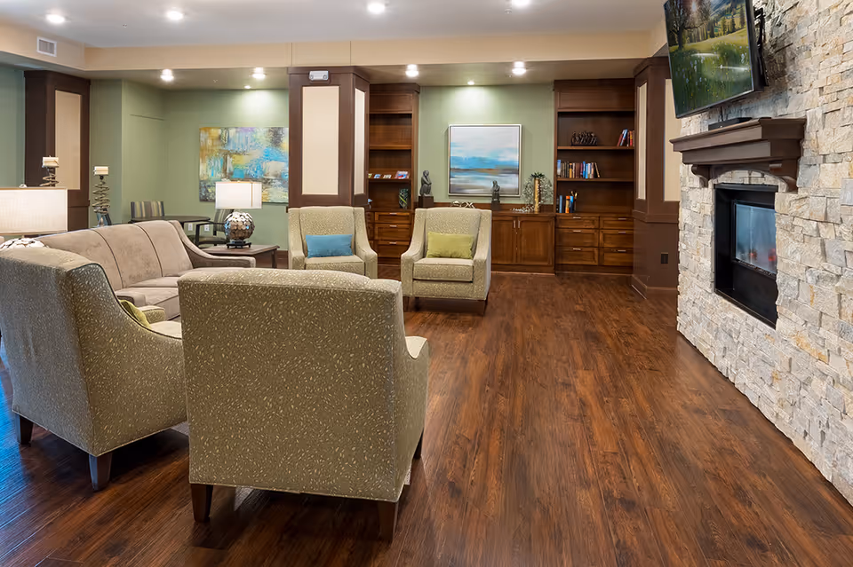 Warm, well-lit common living room with upholstered chairs and sofa, wooden floors, built-in bookshelves, and a stone fireplace with wall-mounted TV.