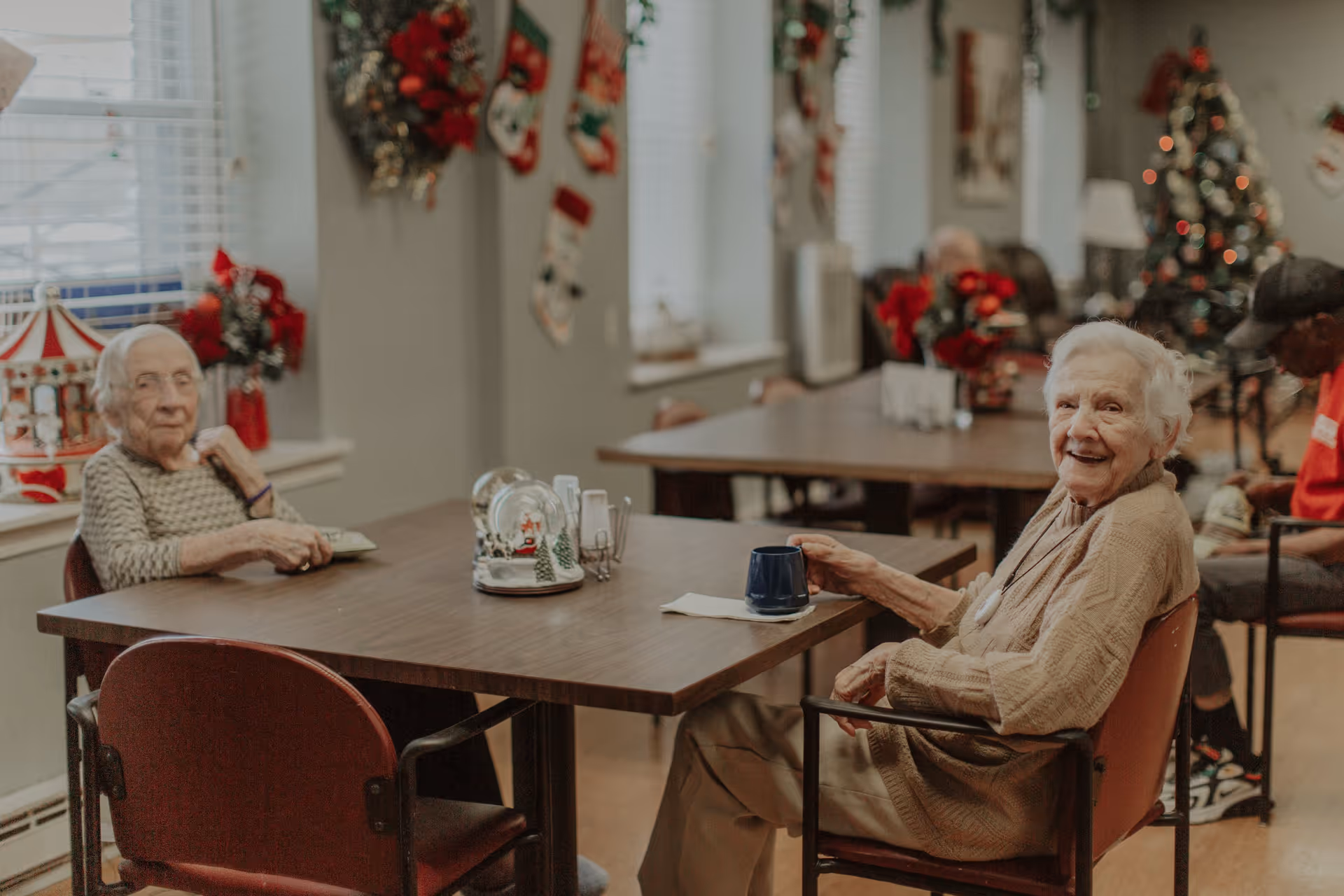 Two elderly women sitting at a wooden table in a decorated room with Christmas wreaths, stockings, and a Christmas tree in the background. One woman is smiling and holding a blue mug, while the other looks toward the camera. Other elderly individuals are seated at tables in the background.