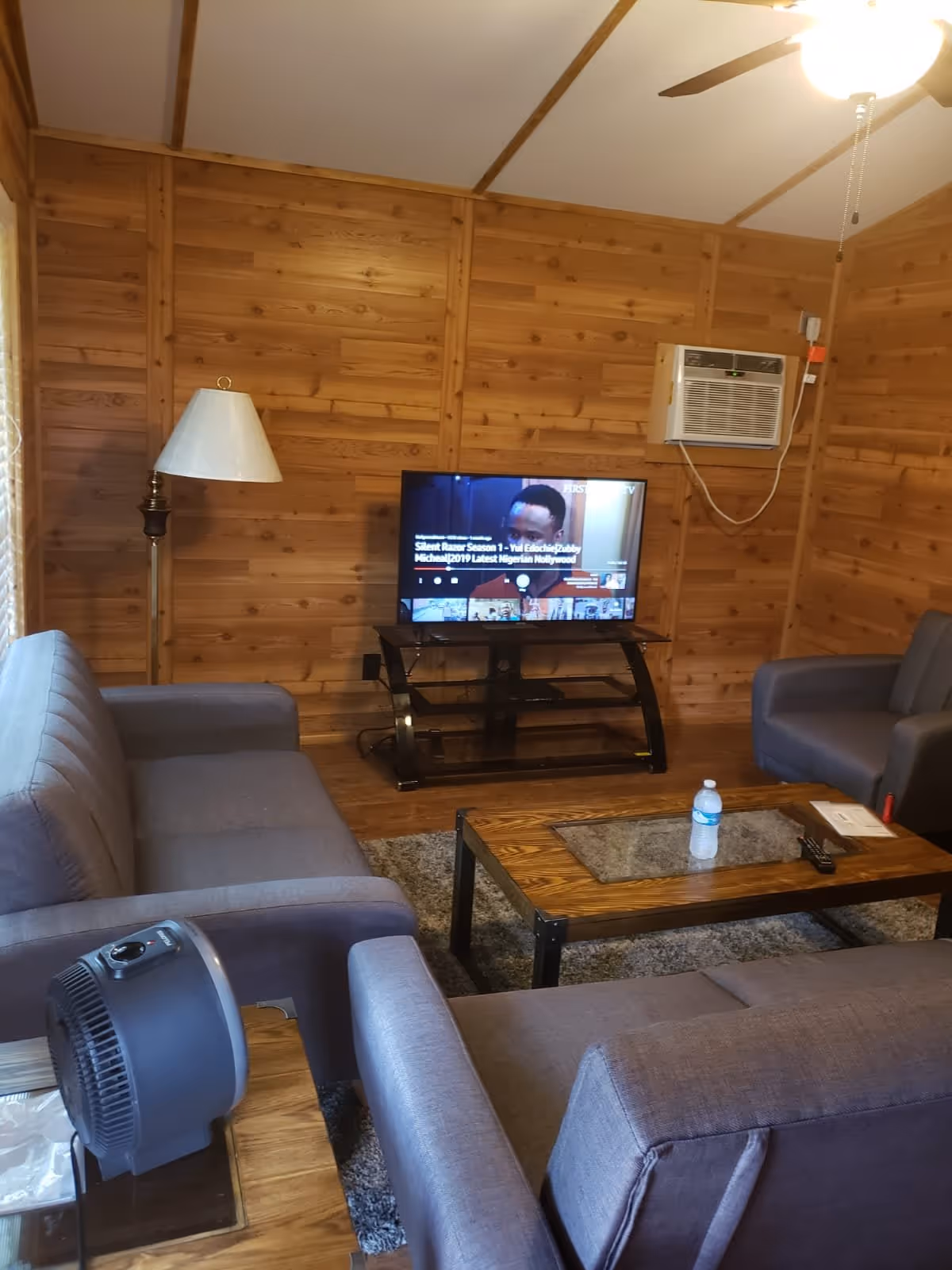 Cozy living room with wooden paneled walls, featuring a flat-screen TV on a black stand, two gray sofas, a wooden coffee table with a water bottle and remote control, a floor lamp, a ceiling fan with light, and a window air conditioning unit.