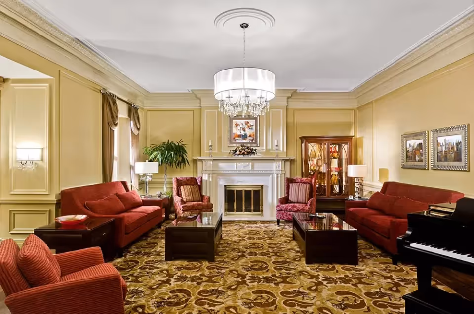 A spacious and elegantly decorated living room with beige walls and a patterned carpet. The room features two red sofas, two red armchairs, a white fireplace with a painting above it, a glass-fronted wooden cabinet, two coffee tables, table lamps, and a black grand piano on the right side.