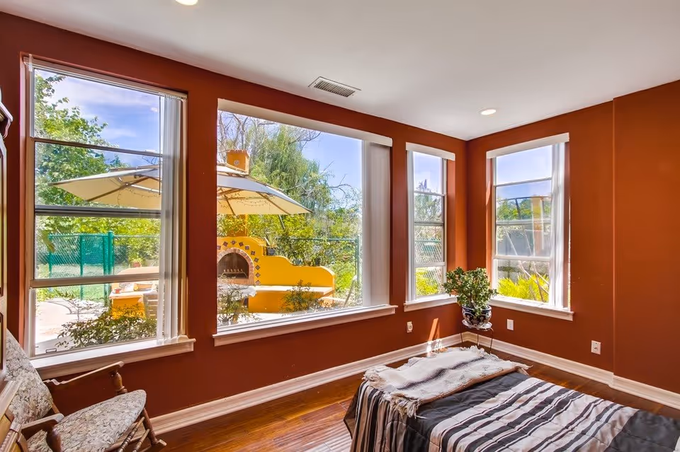 A cozy room with reddish-brown walls and large windows showing an outdoor patio with umbrellas and greenery. Inside the room, there is a striped bed or bench with a blanket and a floral rocking chair. A potted plant is placed near the corner window.