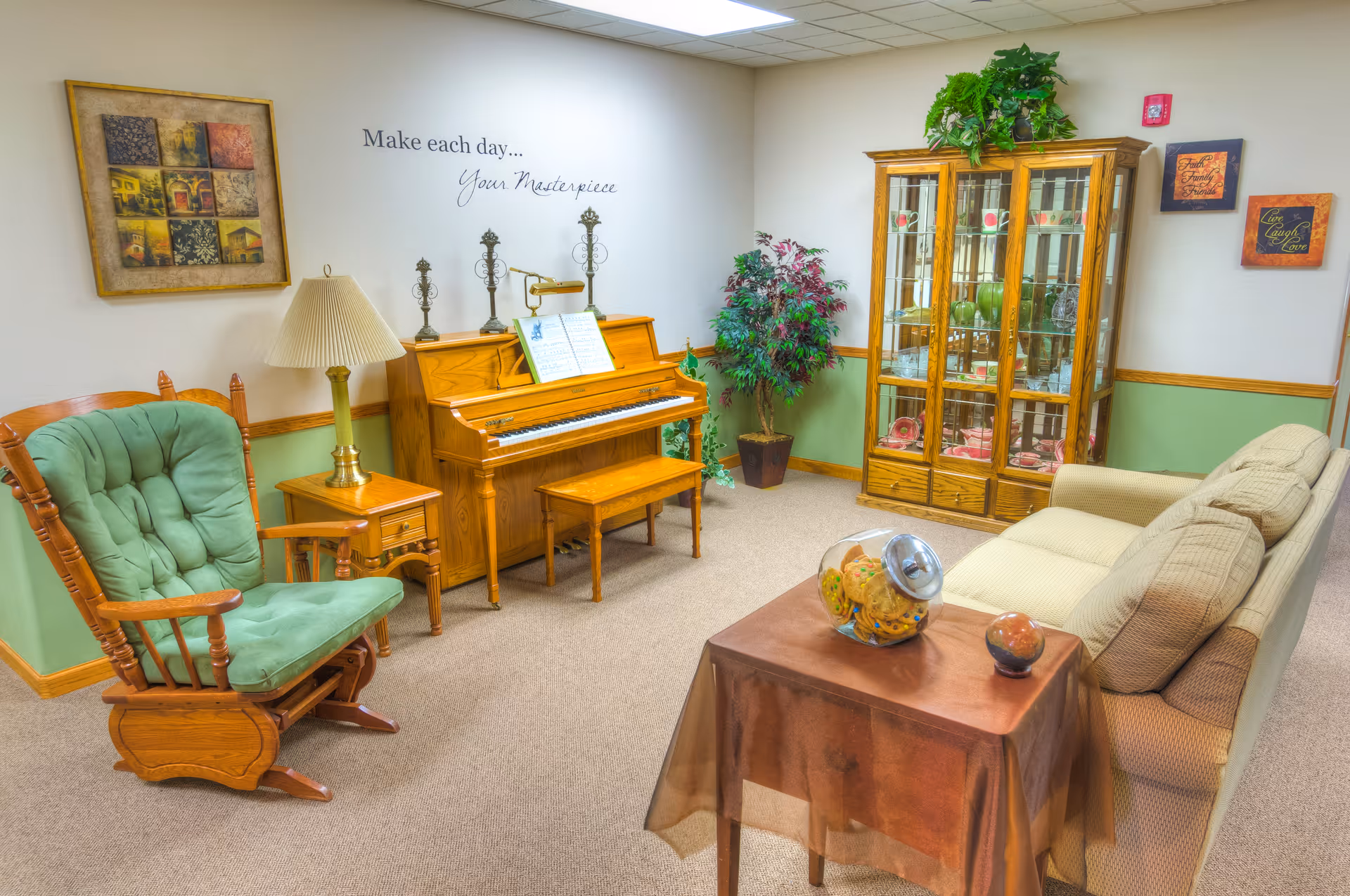 A cozy living room area featuring a wooden piano with sheet music and decorative candle holders on top, a green cushioned wooden rocking chair, a beige sofa, a wooden side table with a lamp, and a glass-front wooden cabinet displaying decorative items. The walls are painted white with a green lower half and adorned with framed artwork and inspirational quotes, including one that says 'Make each day... Your Masterpiece'. There is also a potted plant in the corner.