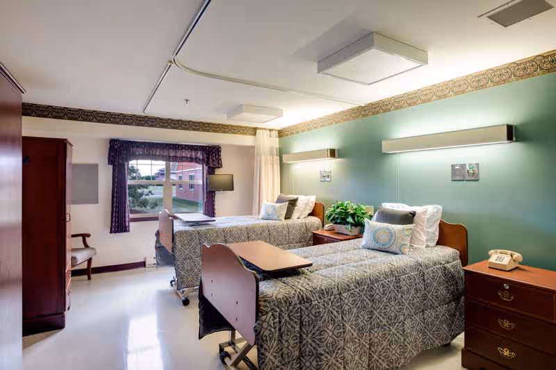 A bright and clean room with two hospital-style beds, each with patterned bedspreads and pillows. There is a wooden nightstand with a telephone and a plant between the beds. The walls are painted green with a decorative border near the ceiling. A window with purple curtains lets in natural light, and there is a wooden chair and a wardrobe on the left side of the room.