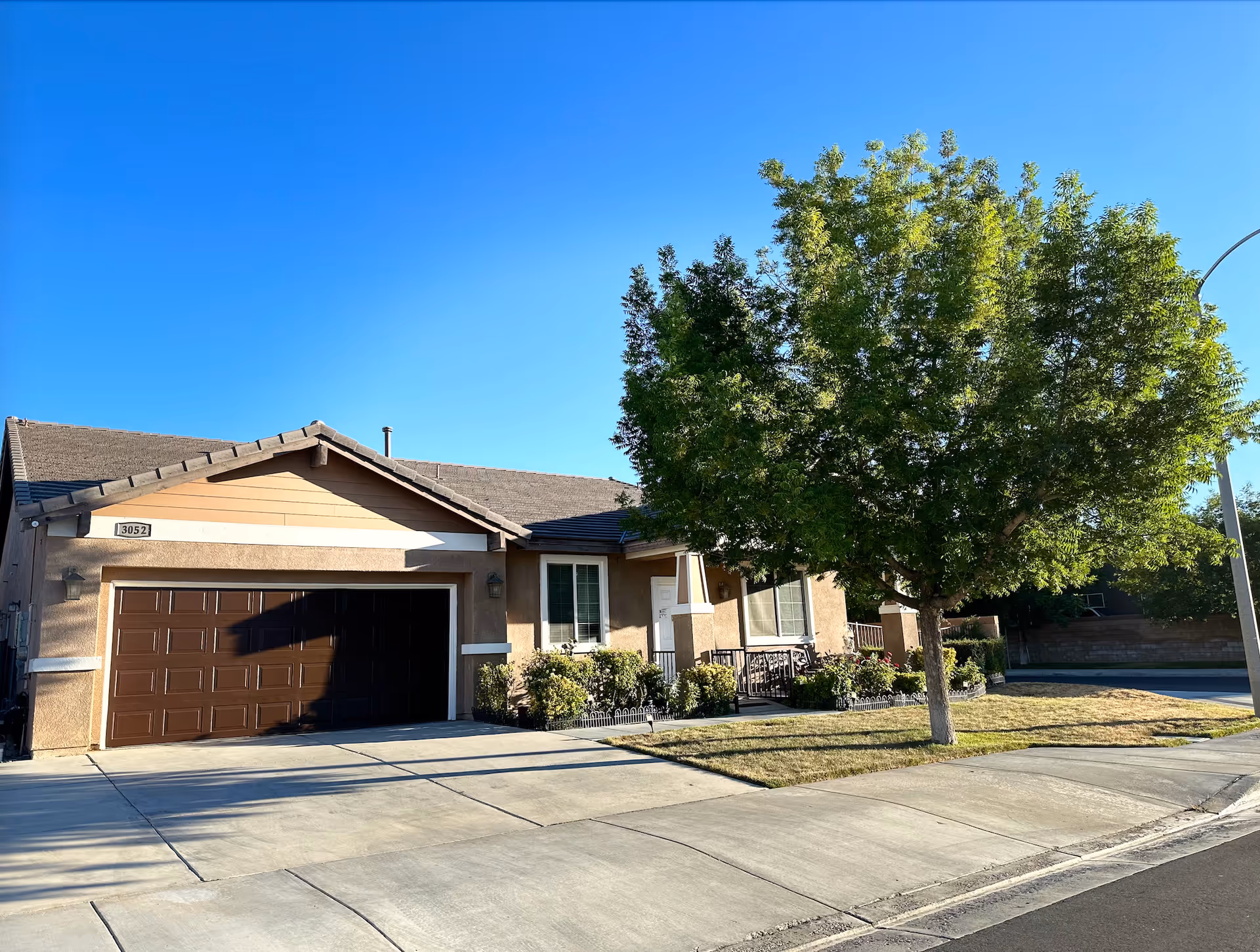 Exterior view of a single-story residential building with a brown garage door, beige walls, and a sloped roof. There is a tree and some bushes in the front yard, and a clear blue sky overhead.