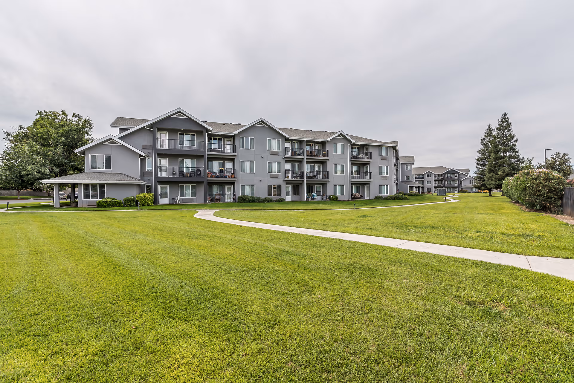 Exterior view of a multi-story senior living facility building with balconies, surrounded by a large, well-maintained grassy lawn and a paved walkway under a cloudy sky.