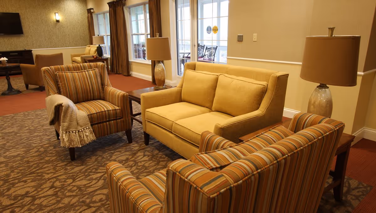A cozy seating area in a senior living facility with a yellow loveseat, two striped armchairs, side tables with lamps, and large windows with brown curtains letting in natural light. The room has warm-toned carpeting and walls, and a door leading to an outdoor patio with chairs is visible in the background.