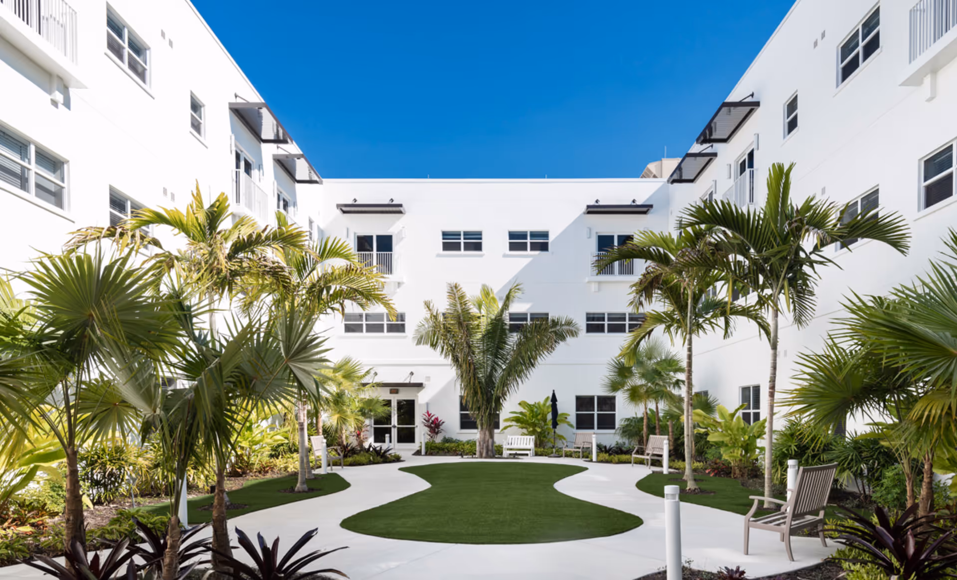 A bright outdoor courtyard area at Amavida Living - Water's Edge featuring a white multi-story building surrounding a landscaped garden with palm trees, green grass, and several benches along a curved white pathway under a clear blue sky.