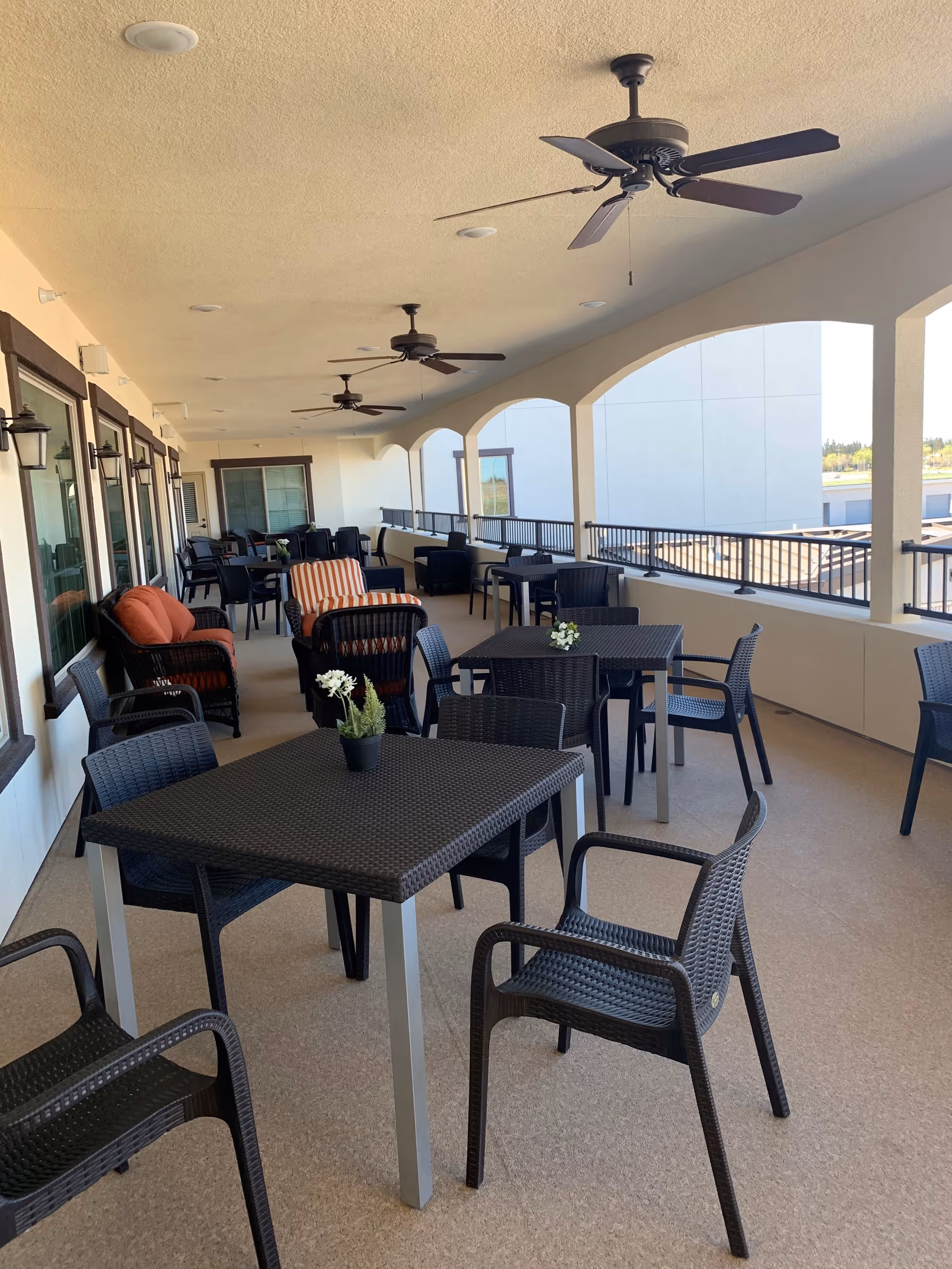 Covered outdoor patio area with multiple black tables and chairs arranged for seating. Ceiling fans are mounted on the ceiling, and there are cushioned lounge chairs with orange and striped pillows along the wall with windows. The patio has arched openings with railings overlooking an exterior view.