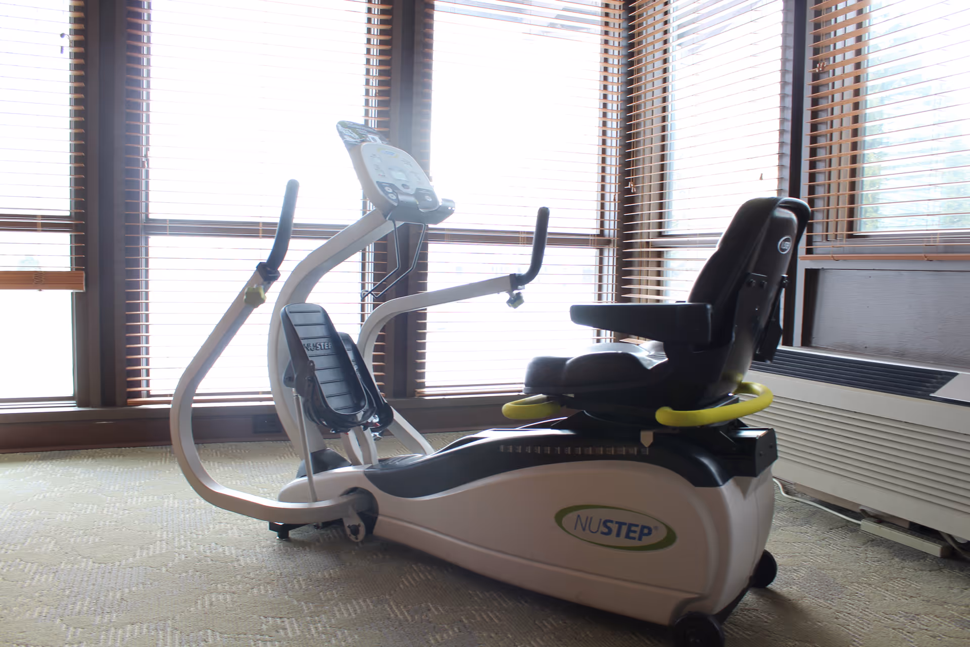 A NuStep recumbent cross-trainer exercise machine positioned in front of large windows with wooden blinds.