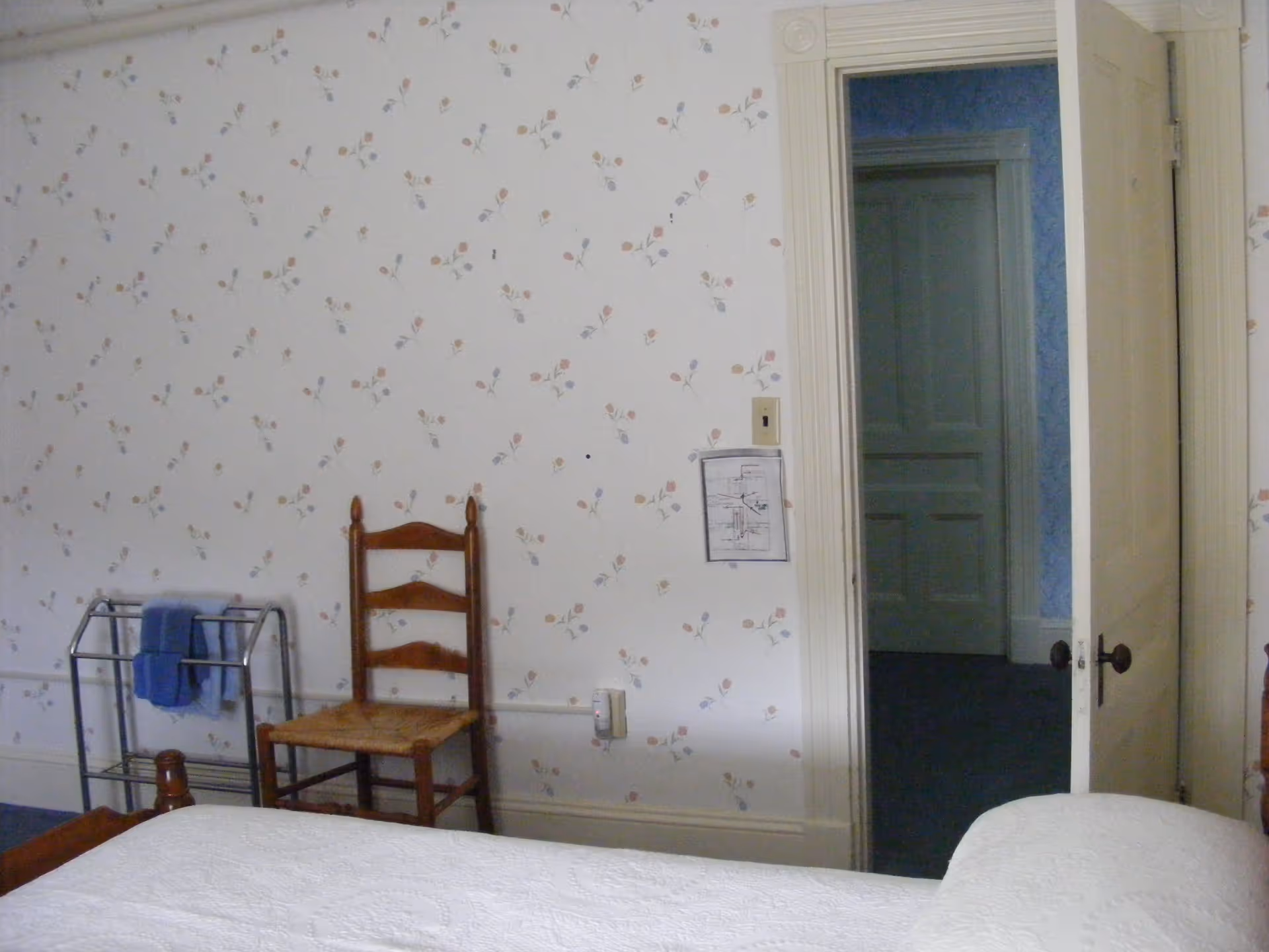 A bedroom with floral wallpaper, a wooden chair with a woven seat, a metal towel rack holding blue towels, and a partially open door revealing a hallway with blue wallpaper and a closed door.
