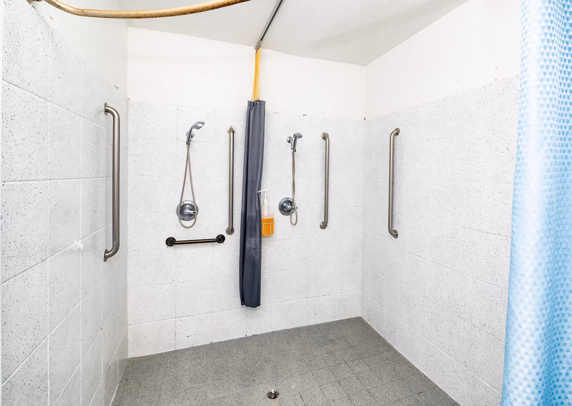 A tiled shower area with two handheld showerheads, multiple stainless steel grab bars on the walls, a gray shower curtain dividing the space, and a bottle of orange liquid soap hanging on one of the showerheads.