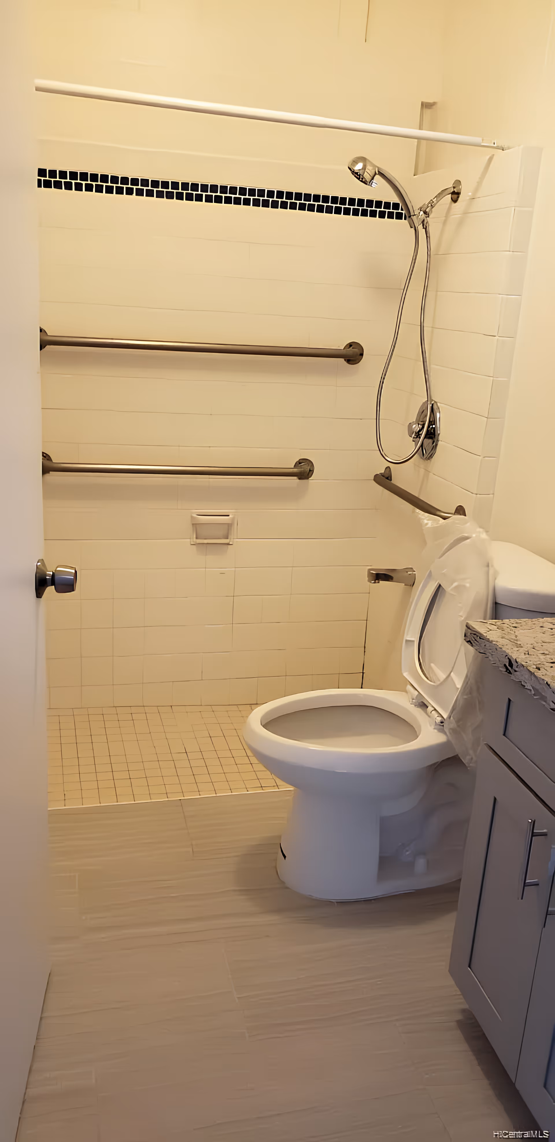 A bathroom with a walk-in shower featuring a handheld showerhead and three horizontal grab bars on the tiled wall. There is a toilet with the seat lid open next to a granite countertop and a cabinet. The floor is tiled in a light color.