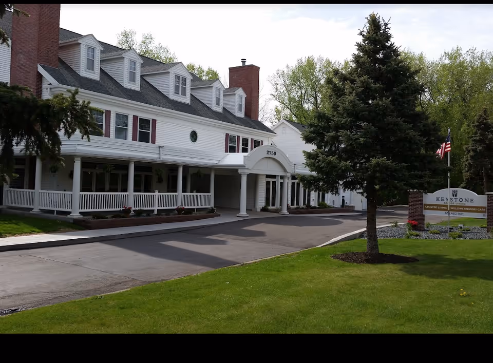 Front exterior of a white multi-story senior living building with a covered entrance, driveway, lawn, and a sign near the road.
