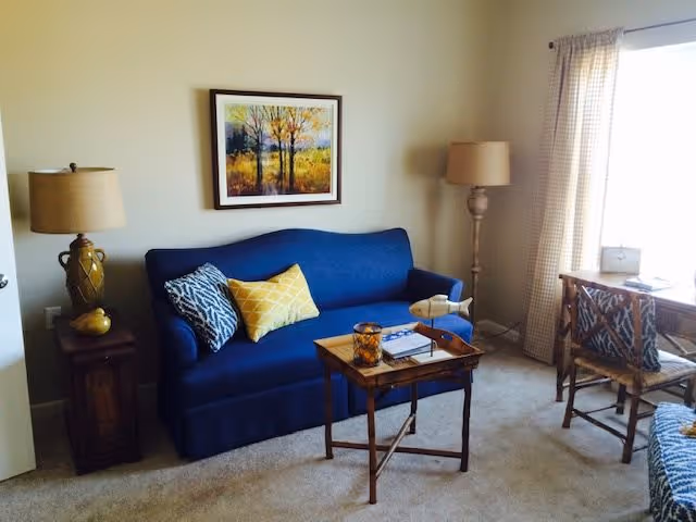 A cozy living room with a blue sofa adorned with two decorative pillows, one yellow and one patterned. A wooden side table with a lamp and a small decorative item is to the left of the sofa. A wooden coffee table in front of the sofa holds a candle, a book, and a small decorative airplane. To the right, there is a wooden desk with a chair near a window with light-colored curtains. A floor lamp stands in the corner near the window, and a framed painting of trees hangs on the wall above the sofa.
