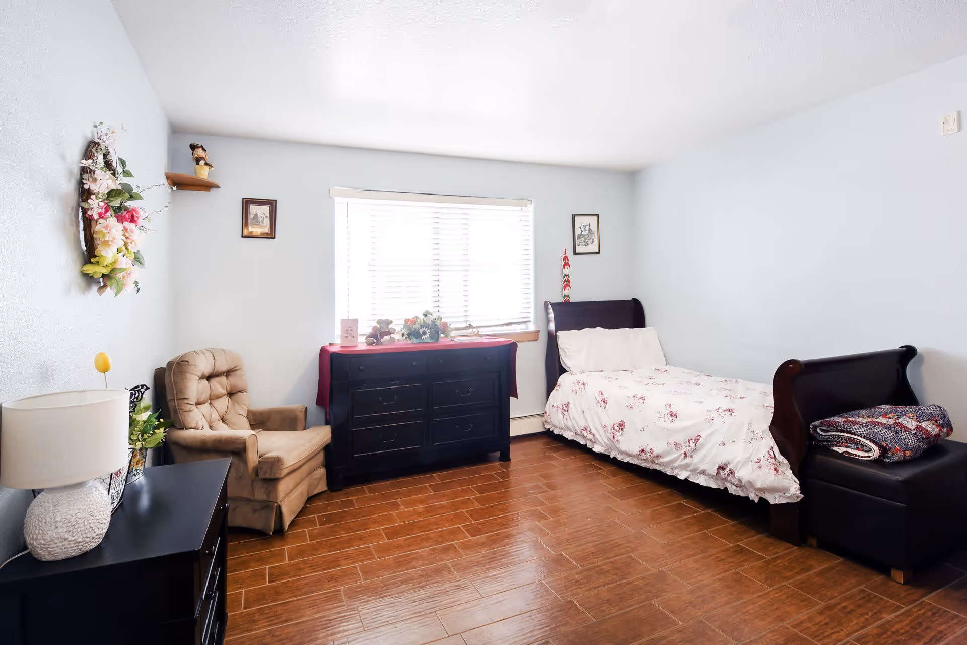 Bright single-occupancy bedroom with a bed, dark dresser under a window, an armchair, and wood-look flooring.