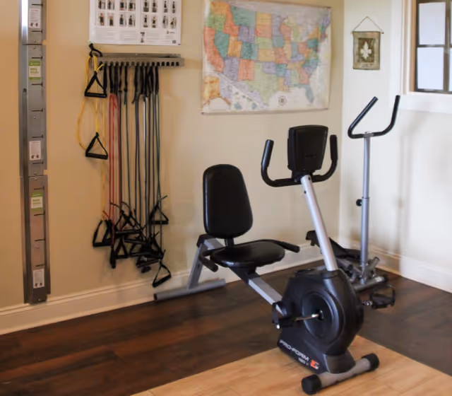 Indoor exercise room with a recumbent stationary bike and an upright stationary bike on a wooden floor. Resistance bands with handles are hanging on the wall next to a map of the United States.