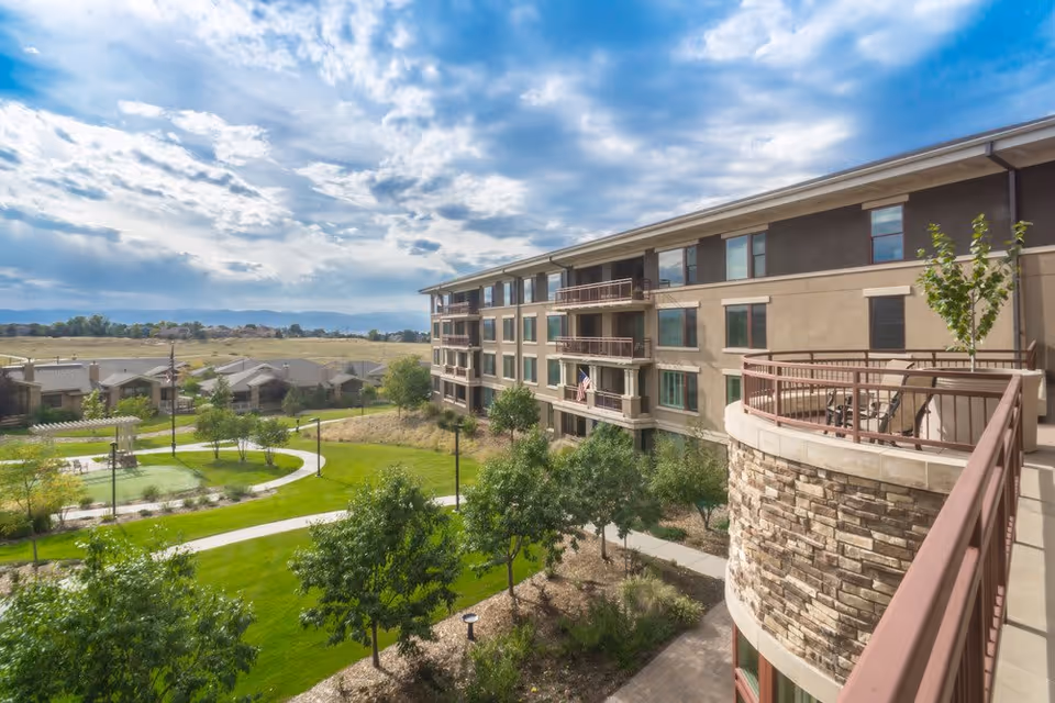 View of a senior living facility building with multiple floors and balconies overlooking a landscaped garden with trees, walking paths, and a pergola under a partly cloudy sky.