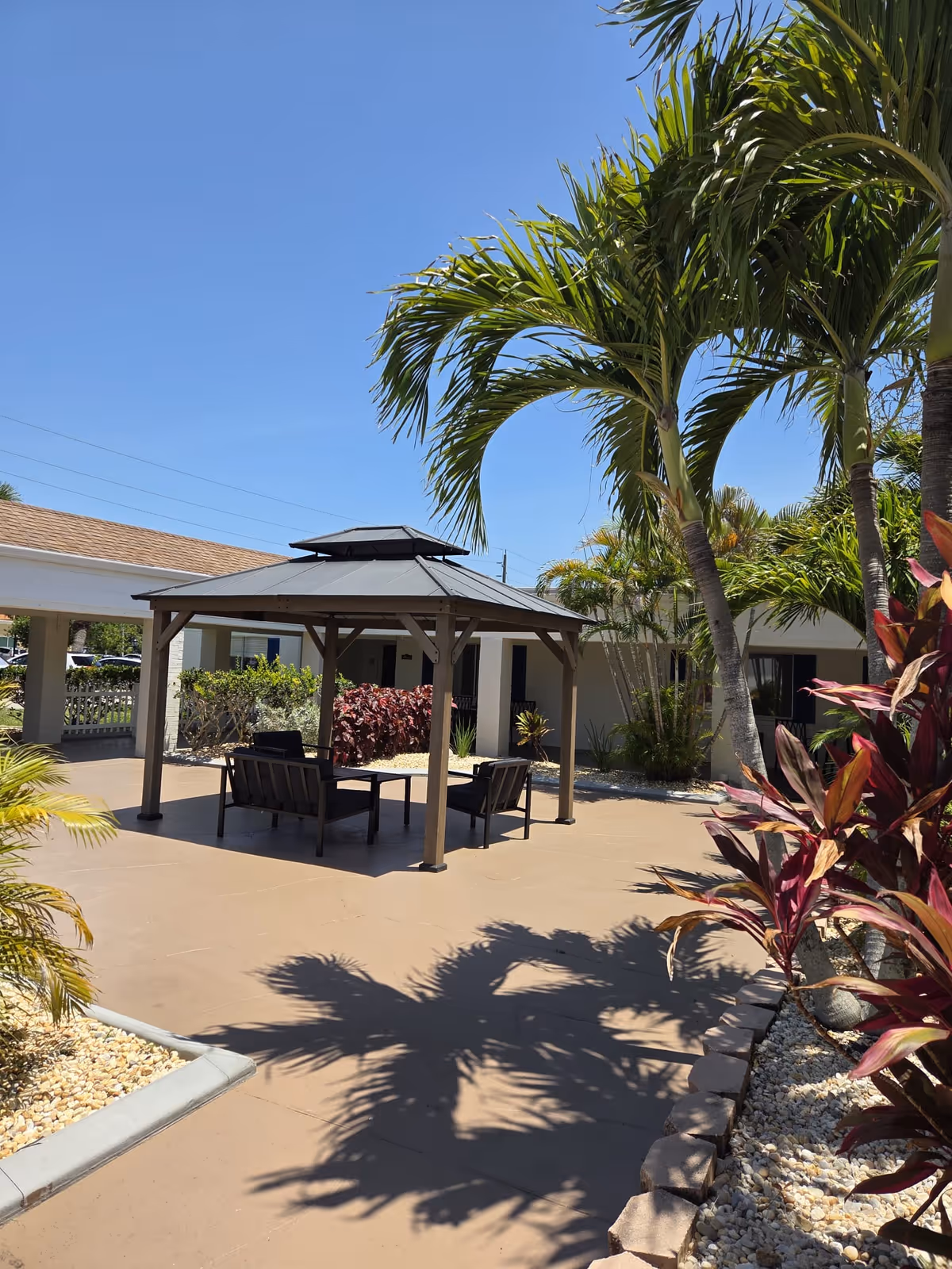 Outdoor patio area at Charlotte Bay Rehabilitation & Care Center featuring a shaded gazebo with seating, surrounded by palm trees and tropical plants under a clear blue sky.