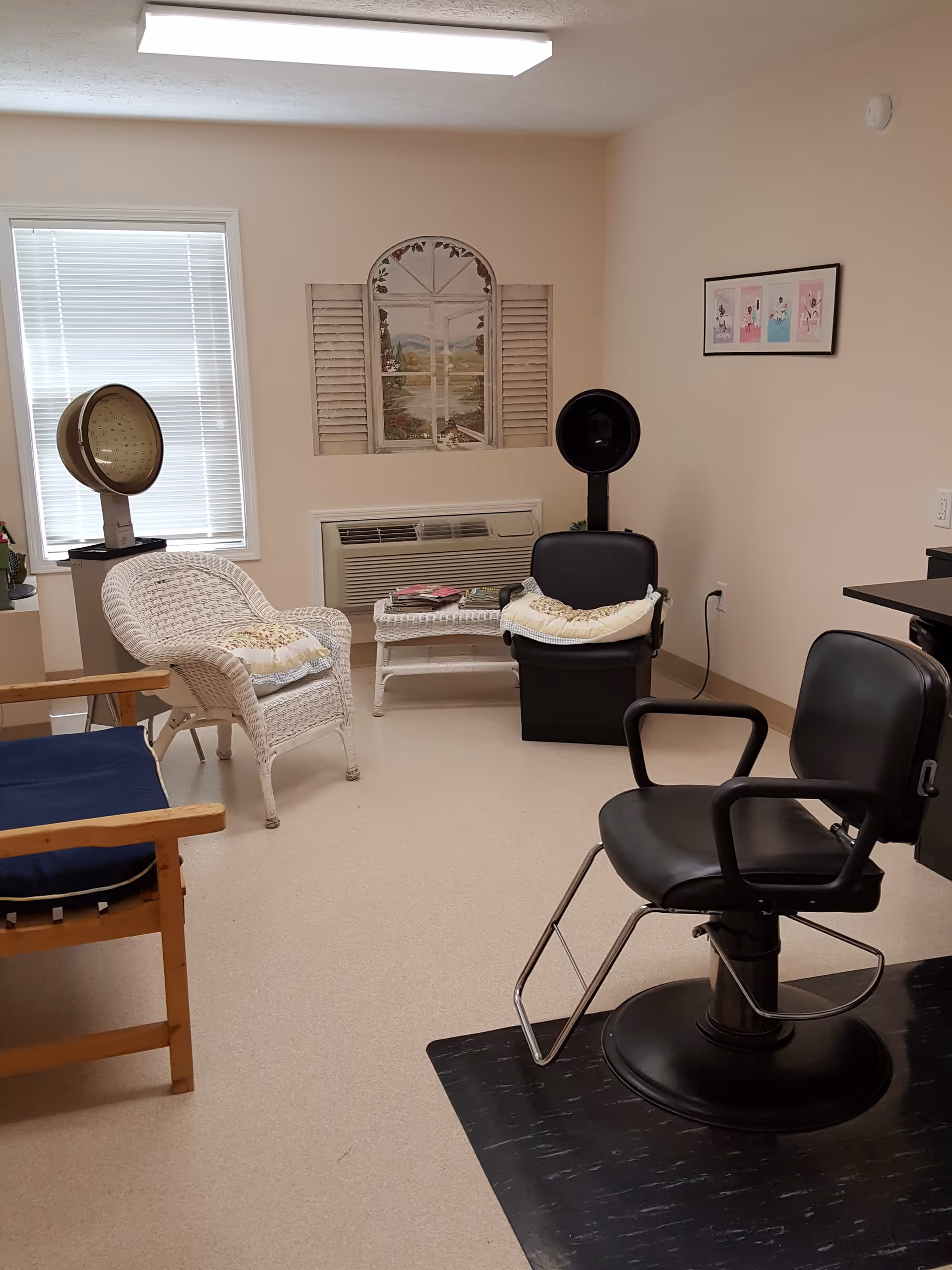 A small salon room with a black salon chair on a black mat, a white wicker chair with a cushion, a wooden chair with a blue cushion, and a black hair dryer chair. There is a window with blinds, a wall-mounted air conditioning unit, and wall decorations including a framed picture and a mural of a window with shutters showing a landscape.