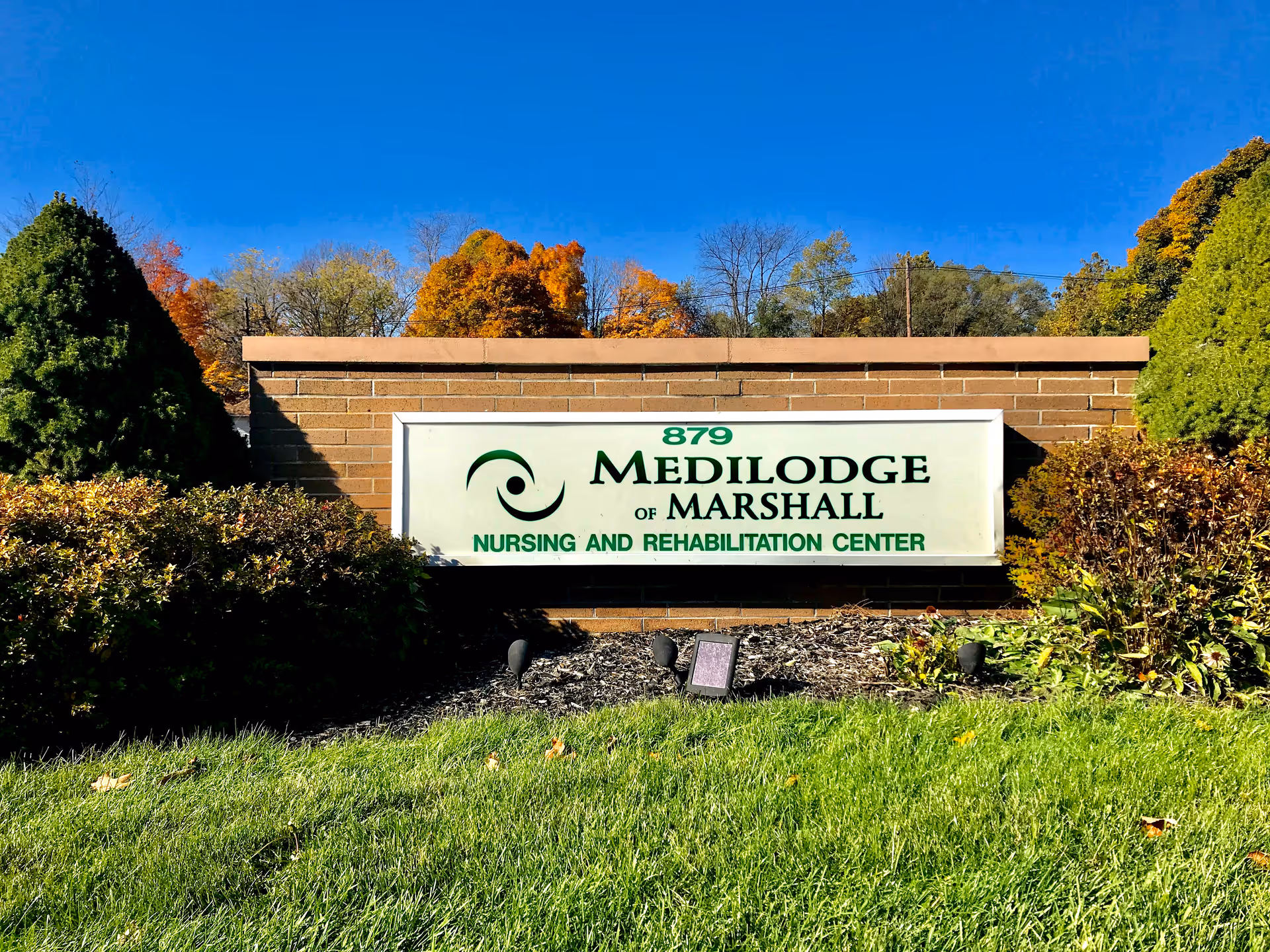 Entrance sign for Medilodge of Marshall nursing and rehabilitation center on a brick wall surrounded by shrubs under a clear blue sky.
