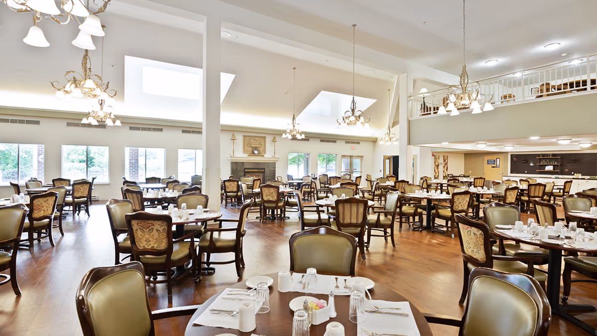 A spacious and well-lit dining room with multiple round tables set with white tableware, glasses, and napkins. The room features wooden floors, elegant chandeliers hanging from a high ceiling, large windows letting in natural light, and a fireplace in the background. There is a mezzanine level with additional seating visible above.