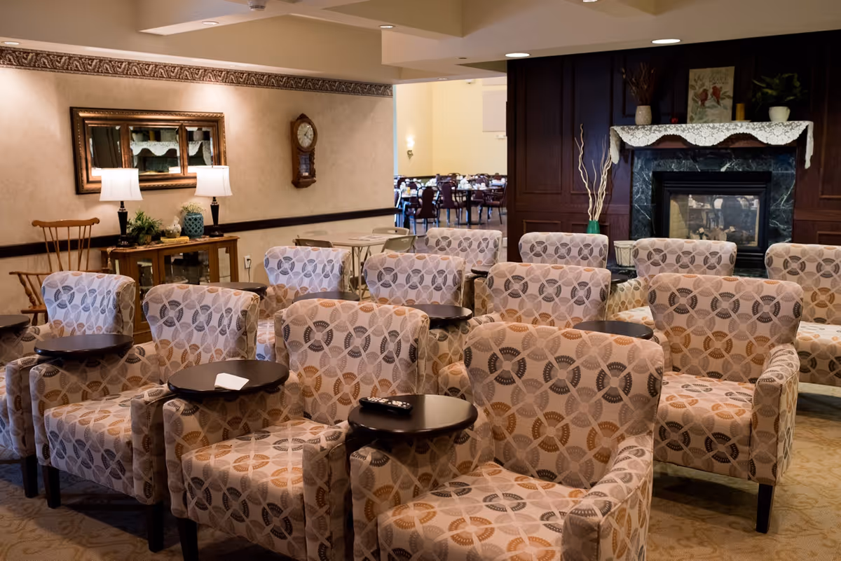 A senior living lounge with patterned armchairs arranged in rows around small side tables, a fireplace and lamps, with a dining area visible in the background.