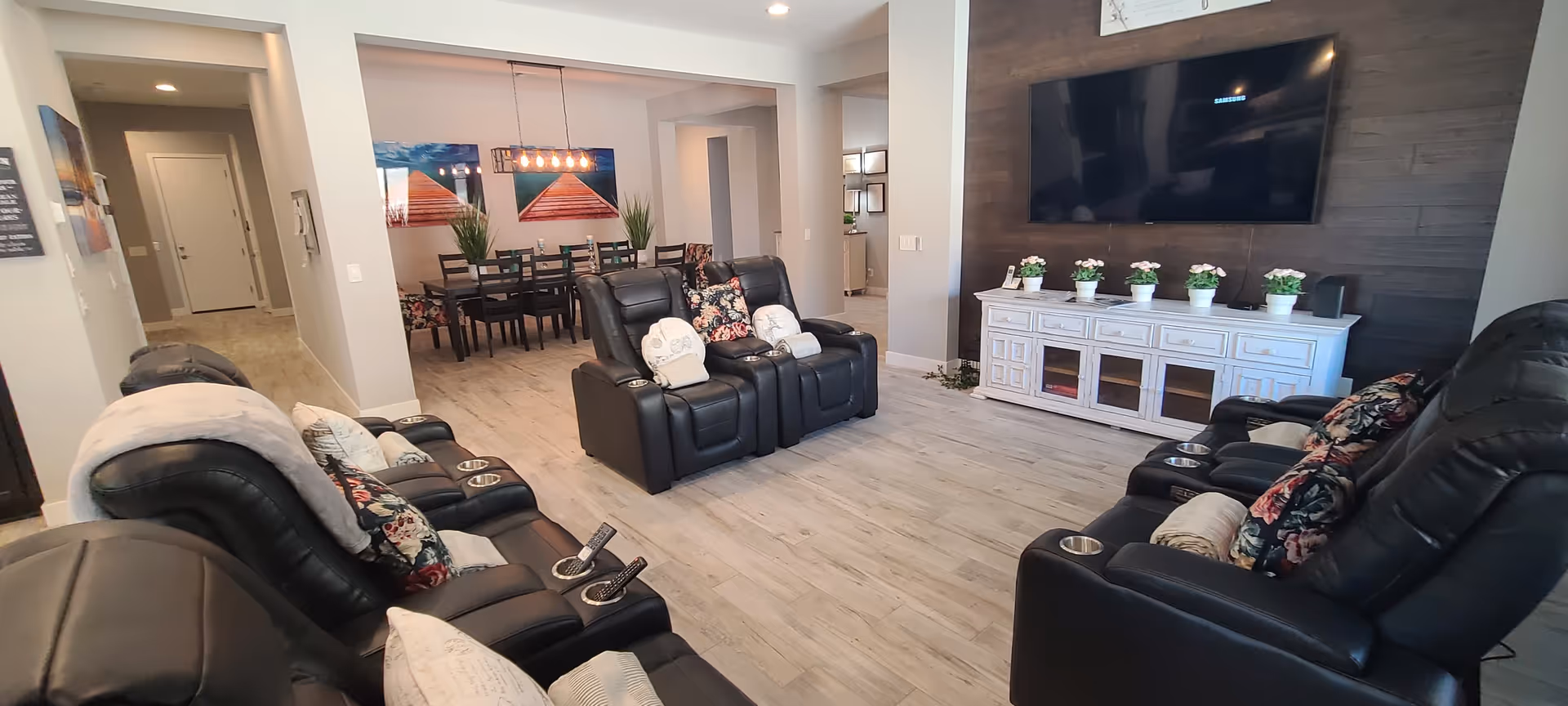 A spacious living room with black leather recliner chairs arranged facing a large flat-screen TV mounted on a dark wood accent wall. A white TV console with decorative potted plants is below the TV. In the background, there is a dining area with a dark wooden dining table and chairs, decorated with two large paintings and green plants. The flooring is light wood, and the walls are painted in neutral tones.