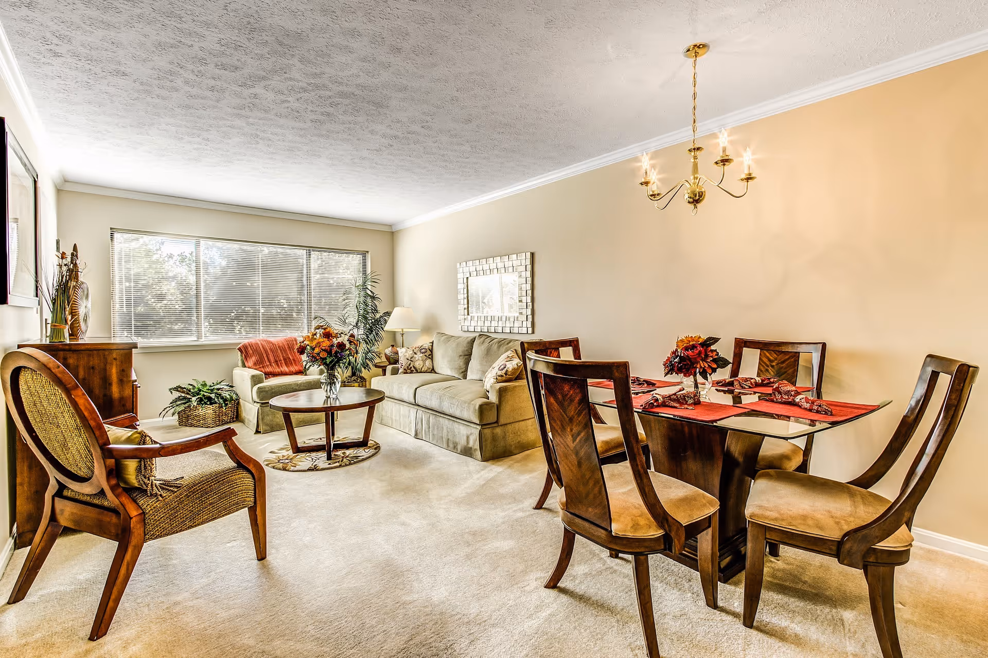 A bright living and dining area with beige walls and carpet. The living room features a beige sofa with patterned cushions, a round wooden coffee table with a floral arrangement, a wooden armchair with woven seat, and a large window with blinds letting in natural light. The dining area has a glass-top table with four wooden chairs and a chandelier hanging above. Decorative plants and framed artwork complete the cozy setting.