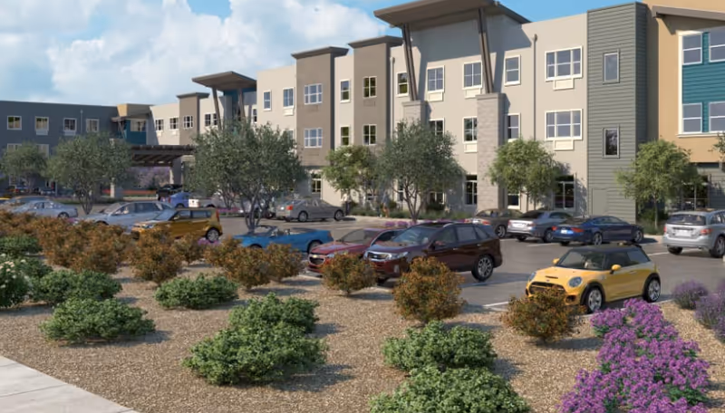 Exterior view of a multi-story senior living facility with a parking lot in front. Several cars are parked, and there are landscaped bushes and trees around the parking area. The building has multiple windows and a modern design with neutral colors.
