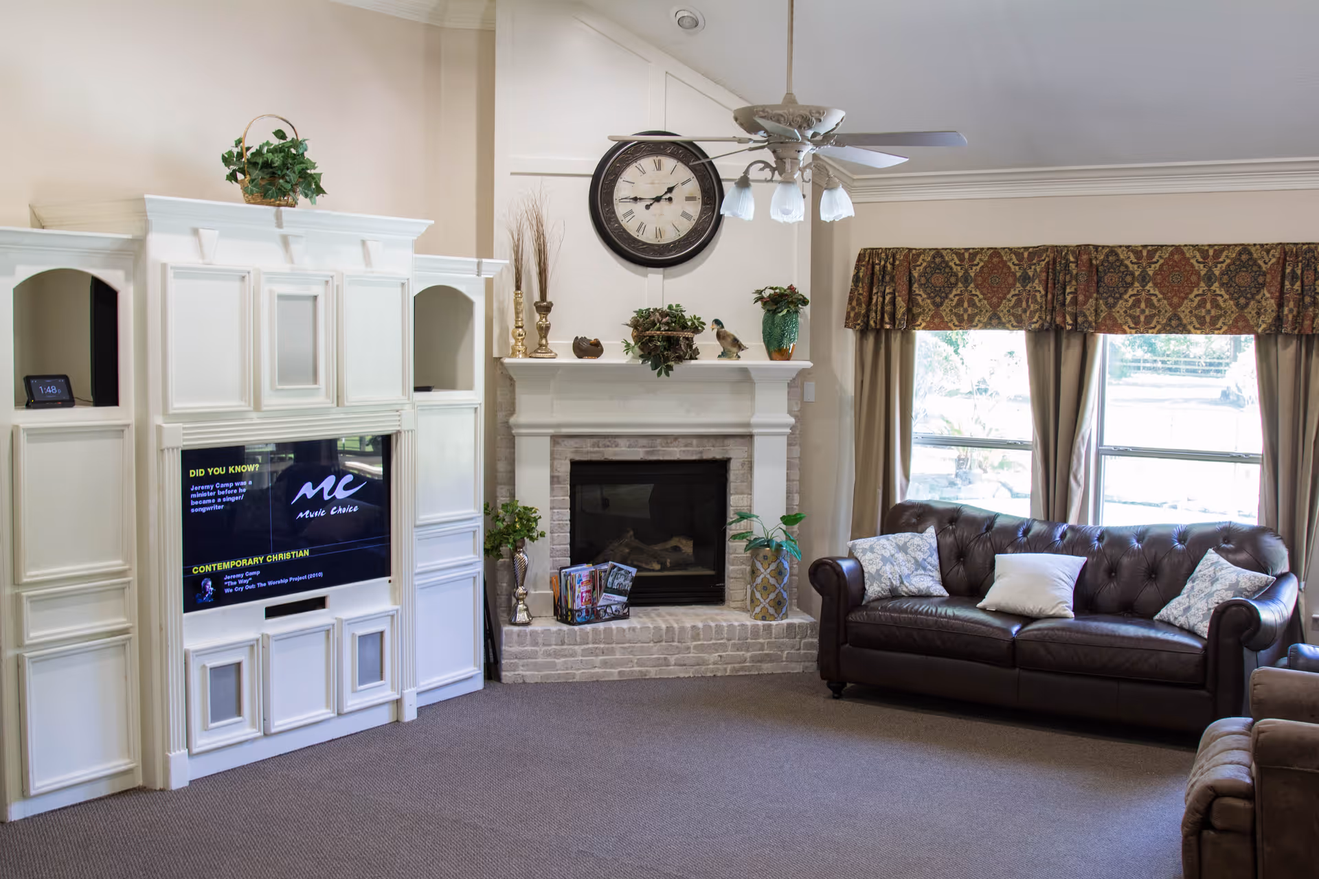 A cozy living room featuring a white entertainment center with a flat-screen TV, a brick fireplace with decorative items on the mantel, a ceiling fan with lights, and a dark brown leather sofa with patterned pillows in front of two large windows with curtains and valances.