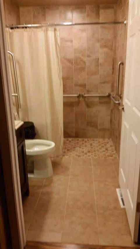 A bathroom with beige tiled floor and walls, featuring a walk-in shower area with grab bars and a shower curtain. There is a white toilet and a dark wooden vanity with a sink on the left side.