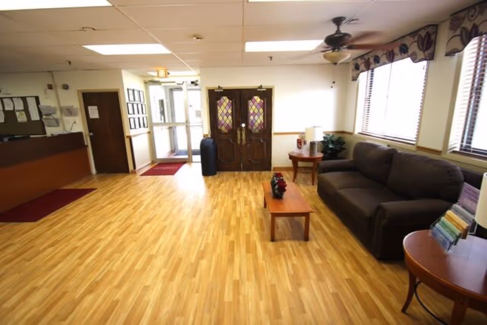 A bright and clean waiting area with wooden flooring, a dark brown couch, two wooden side tables with lamps, a wooden coffee table with a floral centerpiece, and large windows with blinds and valances. There is a ceiling fan above, a reception desk to the left, and double doors with stained glass panels at the back.